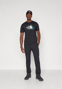 Camiseta negra de algodón con logo en turquesa y blanco, combinada con pantalones negros y zapatos de senderismo negros. Look casual con un diseño ajustado.