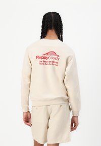 Beige sweatshirt with red graphic text on back; features ribbed cuffs and hem. Paired with matching beige shorts. Minimalist design.