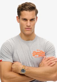 Grijze t-shirt met een oranje "SuperDry" logo, slim fit, korte mouwen, gedragen met een beige leren bandhorloge met een donkere wijzerplaat.