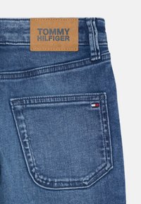 Denim jeans z rjavim usnjenim oznako "TOMMY HILFIGER", modrim pranjem in zadnjim žepom z rdečimi, belimi in modrimi detajli.