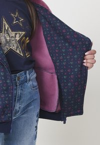 Una persona con una maglietta blu a stelle e jeans blu tiene aperta una giacca navy con fodera in pile rosa e una stampa di piccoli cuori e triangoli.