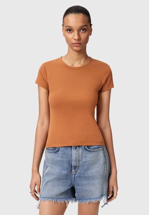 Kvinde med trukket hår tilbage iført en tætsiddende rustfarvet kortærmet skjorte og lyseblå slidte denimshorts mod en grå baggrund.