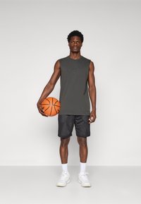 Utenærmet mørkegrå treningstopp med en subtil logo, kombinert med svarte mesh-shortser, holder en oransje basketball, iført hvite joggesko.