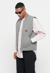 Άνδρας που φορά μαύρα γυαλιά ηλίου, γκρι ζακέτα με φερμουάρ και παραλλαγές, ροζ πουκάμισο κουμπωτό και μαύρο παντελόνι, στέκεται με το ένα χέρι στην τσέπη.