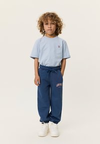 Ανοιχτό γαλάζιο βαμβακερό μπλουζάκι με κοντά μανίκια, συνδυασμένο με μπλε σκούρο αθλητικό παντελόνι με πορτοκαλί τύπωμα. Το σύνολο ολοκληρώνουν λευκά αθλητικά παπούτσια.