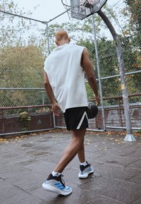 Insieme atletico composto da una canotta bianca senza maniche e pantaloni corti neri con accenti bianchi; indossando scarpe da basket grigie e blu, dribblando un pallone da basket.