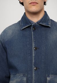 Gros plan d'une personne portant une veste en jean bleue avec des boutons noirs et deux poches avant, col rabattu.