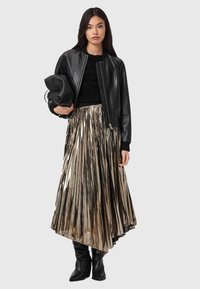 Sort læder bomberjakke over en sort sweater og et plisseret metallisk guldskørt, holdt med en sort taske, parret med knælange sorte støvler.