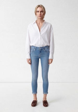 Frau mit blondem Haar in einem weißen Hemd mit Knöpfen, hellblauen hochgekrempelten Skinny-Jeans und braunen Mokassins, die vor einer weißen Wand steht.