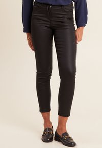 Pantaloni neri, aderenti, realizzati in un materiale lucido, con una vita a media altezza, tasche frontali e una consistenza liscia. Indossati con mocassini neri.