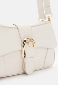 Sac à main de couleur crème avec finition texturée, doté d'une boucle incurvée en or et de fixations de sangle réglables sur fond blanc.