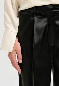 Pantalons en cuir noir présentant une texture lisse, une coupe ajustée et un subtil éclat, associés à une chemise à manches longues de couleur crème.