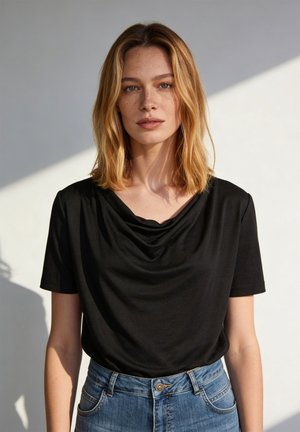 Frau mit schulterlangem blonden Haar, die ein schwarzes T-Shirt mit drapiertem Ausschnitt und blaue Jeans vor hellem Hintergrund trägt.