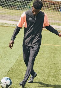 Čierny a ružový športový komplet s bundou na zips a nohavicami, z ľahkého materiálu, nosený osobou na zelenej tráve, futbalová lopta pri jej nohách.