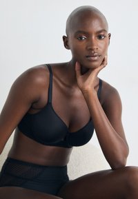 Zwarte onderwired bra met een soepele afwerking, voorzien van verstelbare bandjes en gegolfde randen. Gecombineerd met zwart high-waisted ondergoed met gaasaccenten.