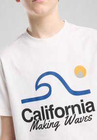 Valkoinen puuvillapaita, jossa on sininen aalto-logo ja musta teksti "California Making Waves". Mukana on myös keltainen ja harmaa auringon graafinen kuva.