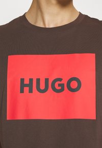 Hnědé bavlněné tričko s velkou červenou obdélníkovou grafikou, ve které je uprostřed tučně napsáno černými písmeny slovo "HUGO".