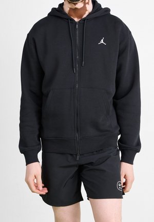 Person trägt einen schwarzen Kapuzenpullover mit Reißverschluss und weißem Jumpman-Logo sowie schwarze Shorts mit Nike Air-Logo am linken Bein.