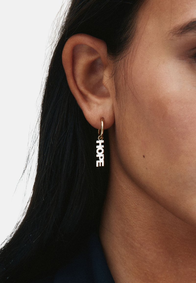 Pendiente de aro de oro con la palabra "HOPE" en letras mayúsculas, con un acabado liso, colgando justo debajo del lóbulo de la oreja.