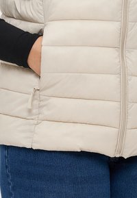 Šviesiai ruda pūkinė liemenė su horizontaliomis siūlėmis, šoniniu užtrauktuku ir priekinėmis užtrauktukais, derinama su tamsiai mėlynais džinsais. Sklandi tekstūra.