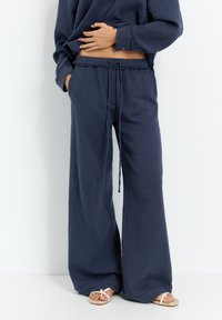 Marineblaue weit geschnittene Hosen aus strukturiertem Stoff mit einem Kordelzugbund. Kombiniert mit weißen Sandalen, die eine entspannte Silhouette zeigen.