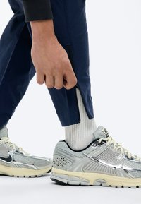 Χέρι που ρυθμίζει το φερμουάρ στο πόδι ενός σκούρου μπλε παντελονιού, φορεμένου πάνω από λευκές κάλτσες και γκρι αθλητικά παπούτσια Nike με μεταλλικές ασημένιες λεπτομέρειες.