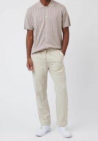 Chemise en tricot beige à manches courtes avec col et fermeture boutonnée, associée à un pantalon en lin beige clair et des baskets blanches.