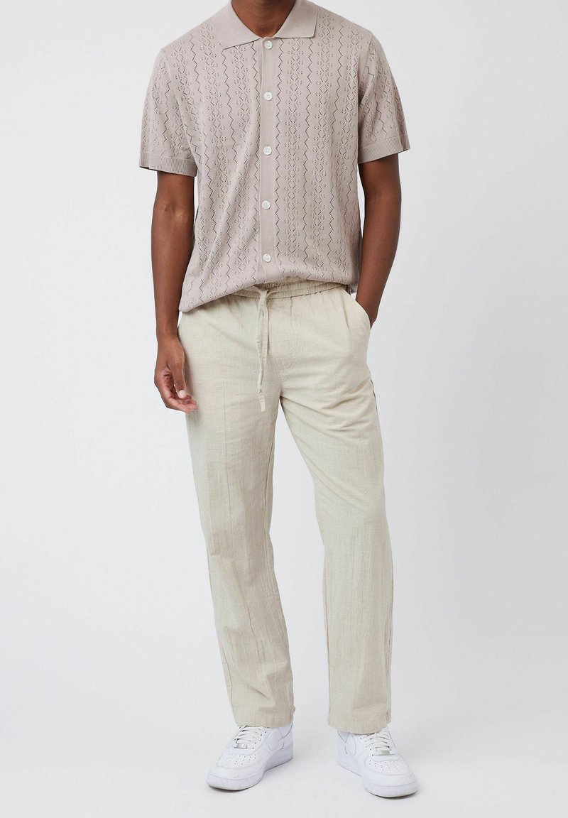 Chemise en tricot beige à manches courtes avec col et fermeture boutonnée, associée à un pantalon en lin beige clair et des baskets blanches.