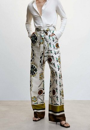 Person trägt hoch taillierte, weit geschnittene Hosen mit bunten Blumen- und geometrischen Mustern, kombiniert mit einem weißen, langärmligen Hemd mit Knöpfen.
