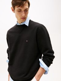 Schwarzer Kapuzenpullover mit Rundhalsausschnitt und gerippten Bündchen, über einem hellblau gestreiften Hemd getragen. Auffälliges kleines Logo auf der Brust.