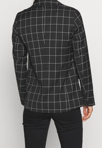 Black blazer featuring a grid pattern in white, tailored fit with a lapel collar, long sleeves, and structured shoulders. Textured fabric.