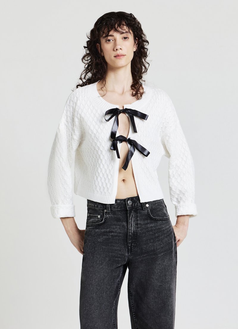 Weißer gesteppter Crop-Pullover mit schwarzen Ripsband-Bindebändern an der Vorderseite, kombiniert mit dunkelblauen, hoch taillierten Jeans. Weicher Stoff und runder Ausschnitt.