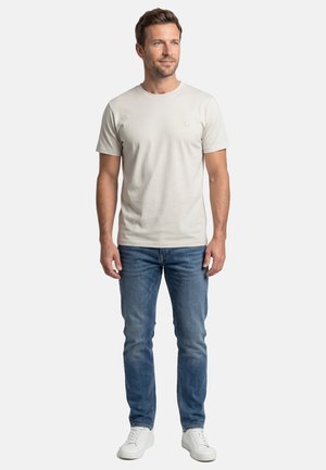 Mann steht, trägt ein schlichtes beige T-Shirt mit kurzen Ärmeln, blaue Jeans und weiße Turnschuhe, mit neutralem Gesichtsausdruck.