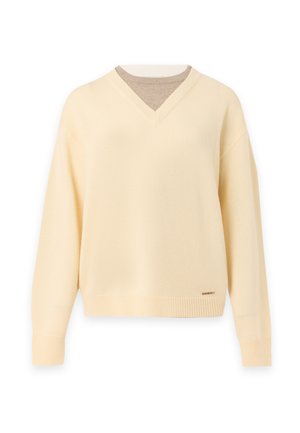 Femme portant un pull beige col en V, une jupe plissée asymétrique noire, des bottes noires, et tenant un sac à main noir sur un fond uni.