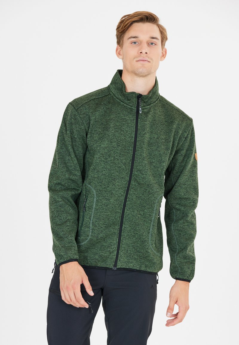 Un giovane che indossa una giacca in pile verde con zip e pantaloni neri, in piedi contro uno sfondo bianco semplice.