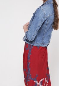 Veste en jean à coupe ajustée, de couleur bleue, avec fermeture à boutons sur le devant, portée par-dessus une robe maxi rouge à fleurs avec des motifs bleus complexes.