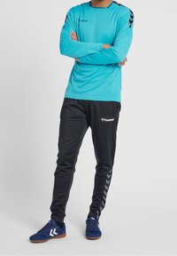 Türkises Langarmhemd mit schwarzen Schulterakzenten, kombiniert mit schwarzen Jogginghosen mit kontrastierenden seitlichen Chevron-Details und blauen Sneakers.