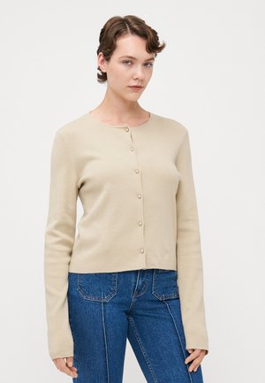 Beige cardigan med knappestenging foran med perlelignende knapper, ribbestruktur, rund halslinje og lange ermer, kombinert med blå jeans.