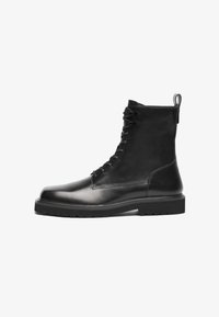 Bota de cuero negra hasta la mitad de la pantorrilla con cordones, puntera lisa, suela gruesa y tira de sujeción sobre un fondo blanco, vista de lado.