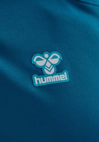 Turquoise sportshirtstof met een gestructureerd oppervlak. Heeft een blauw en wit bijenlogo en "hummel" tekst in een moderne lettertype.