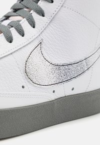 Bílé kožené tenisky se šedými tkaničkami, texturovanou šedou gumovou podešví a charakteristickým logem Swoosh s tečkovaným vzorem.
