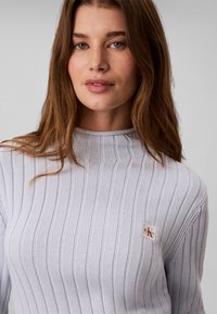 Jeune femme aux longs cheveux bruns portant un pull côtelé gris clair avec un petit écusson logo Calvin Klein sur la poitrine.
