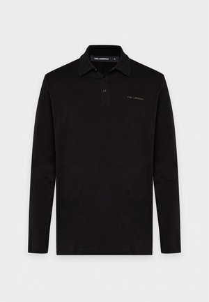 Un homme pose avec un polo noir à manches longues, un pantalon foncé, une casquette noire et des chaussures noires et blanches sur un fond gris clair.