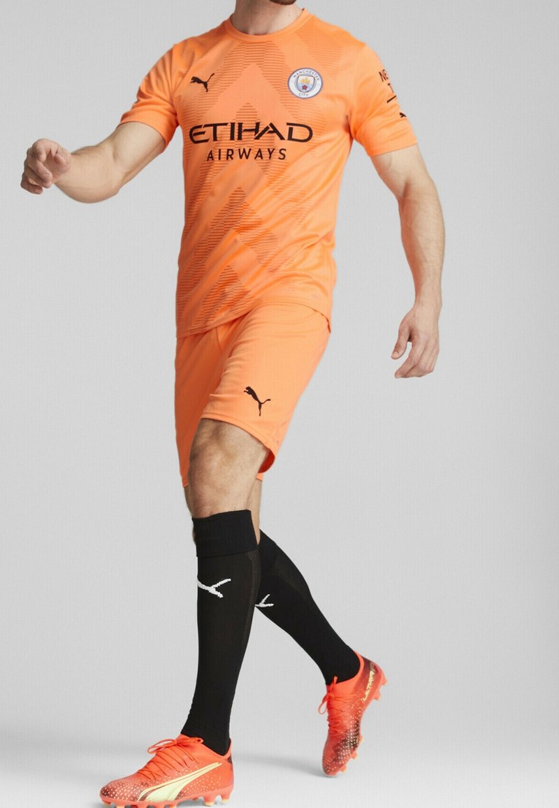 Maglia da calcio arancione e pantaloni corti con loghi neri, abbinati a calze nere e scarpe da calcio arancioni fluorescenti, caratterizzate da motivi testurizzati e dettagli.