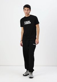 Μαύρο γραφικό T-shirt με λογότυπο "KARL LAGERFELD", συνδυασμένο με μαύρες φόρμες που διαθέτουν λογότυπα. Το μοντέλο φοράει γκρίζα αθλητικά παπούτσια.
