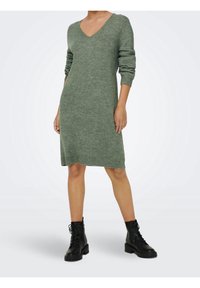 Robe pull en maille verte avec un col en V, des manches longues et des poignets côtelés. Portée avec des bottines noires à lacets. Texture lisse.