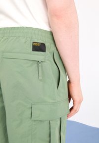 Pantalones cortos de carga verdes con un acabado texturizado, cintura elástica, bolsillos laterales y una etiqueta negra sujeta al bolsillo trasero.