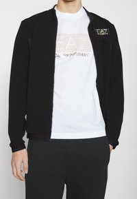 Giacca nera con zip in tessuto testurizzato, caratterizzata da un accento con logo. Indossata sopra una maglietta bianca con un logo stampato in oro e pantaloni scuri.
