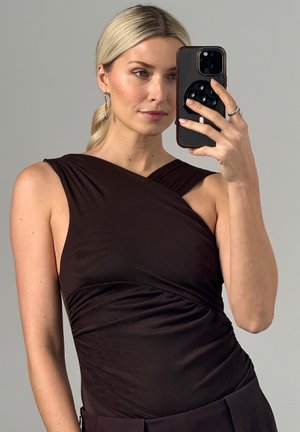 Blondynka w bezrękawniku o asymetrycznym kroju w ciemnym brązowym kolorze robi sobie selfie w lustrze smartfonem na tle jednolitego szarego tła.