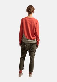 Rode sweatshirt met een ronde zoom en een beige onderpaneel, gecombineerd met kaki cargobroeken met zakken en bandjes, gedragen met lichte schoenen.